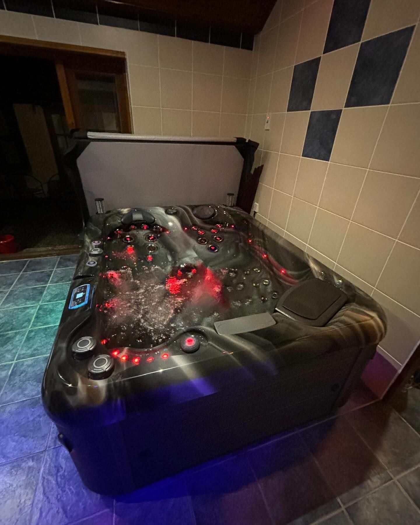 A lit up black hot tub with red and blue lights, partially covered, inside a room with tiled walls.