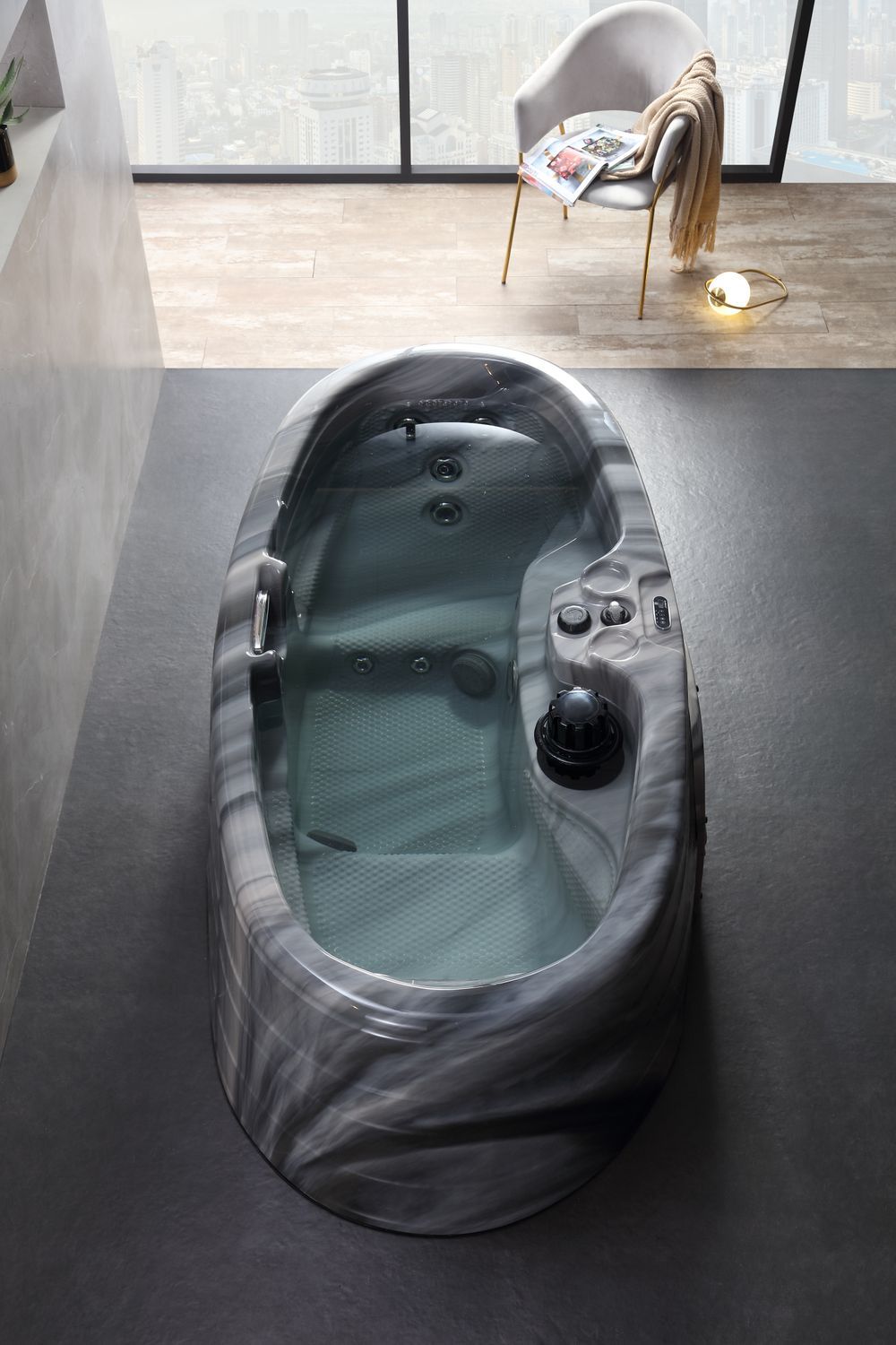 Oval hot tub filled with water, in a modern bathroom setting. Chair with blanket and book near window.