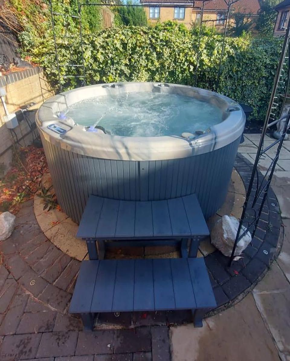 Hot tub in a garden, with blue steps leading up to it. Brick patio and lush green shrubs surround it.