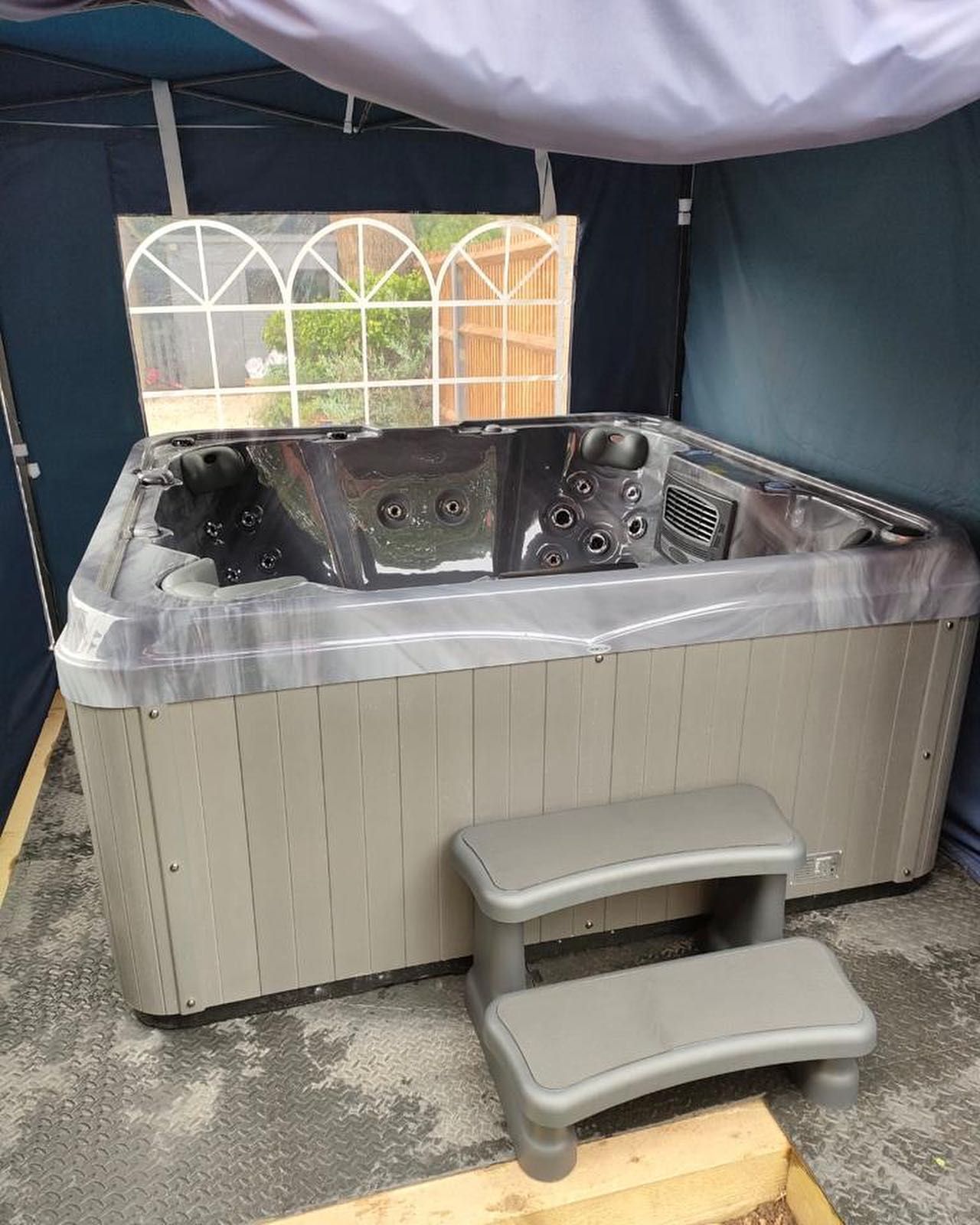 Hot tub with gray siding, two-step gray entry, under a white and blue canopy. Window behind.