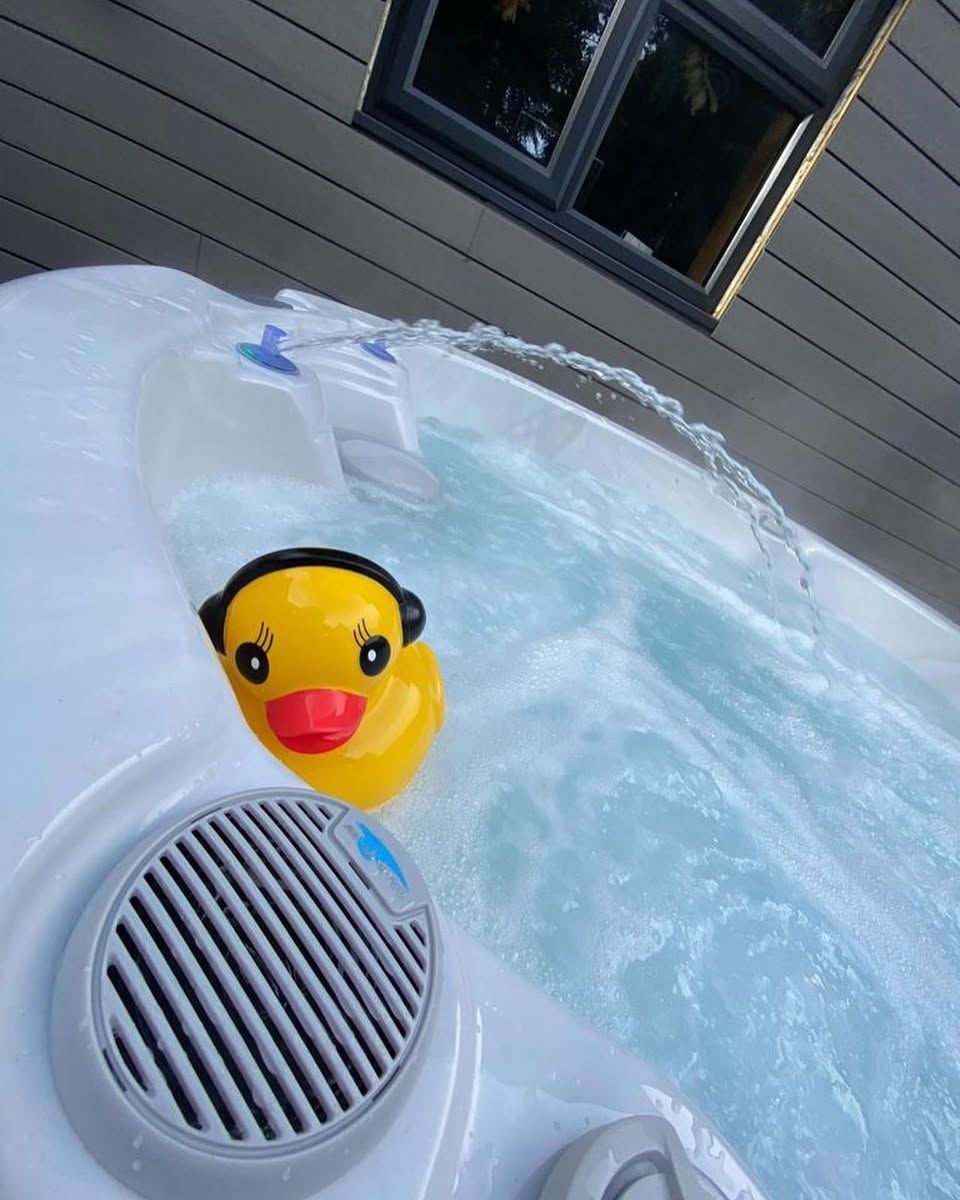 Yellow rubber duck with headphones in a bubbling hot tub next to a speaker, beneath a window.