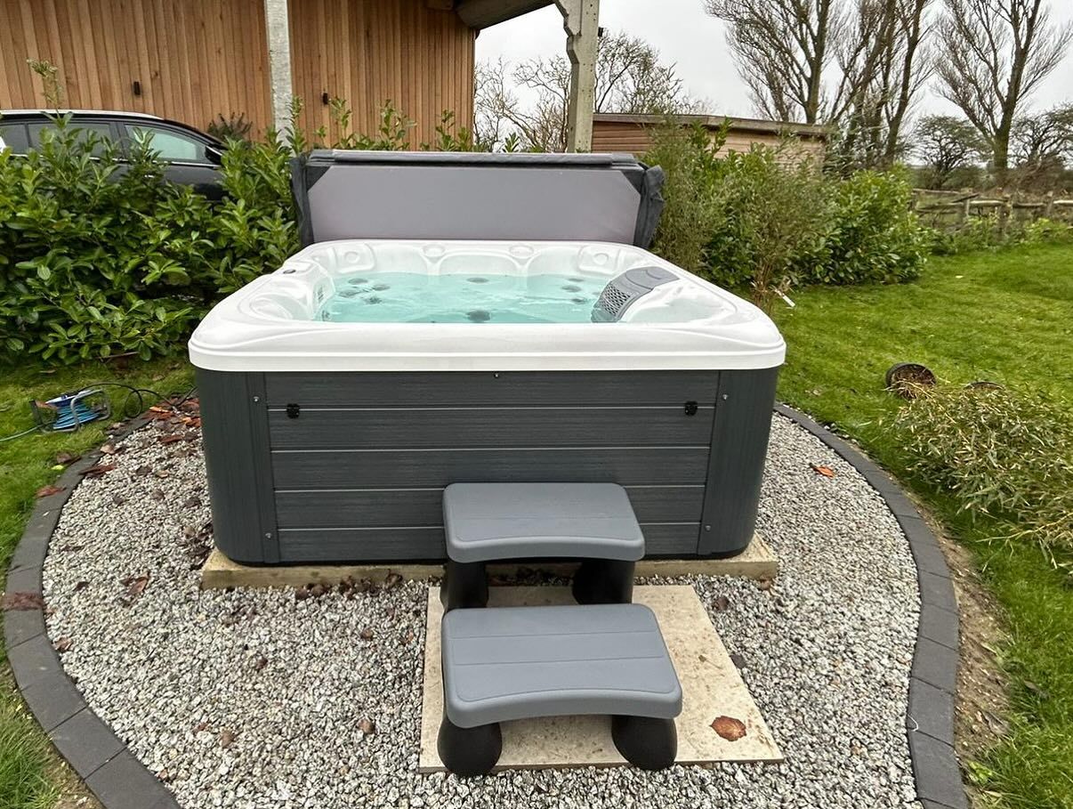 Gray hot tub with partially open cover, on gravel, with steps in a yard.
