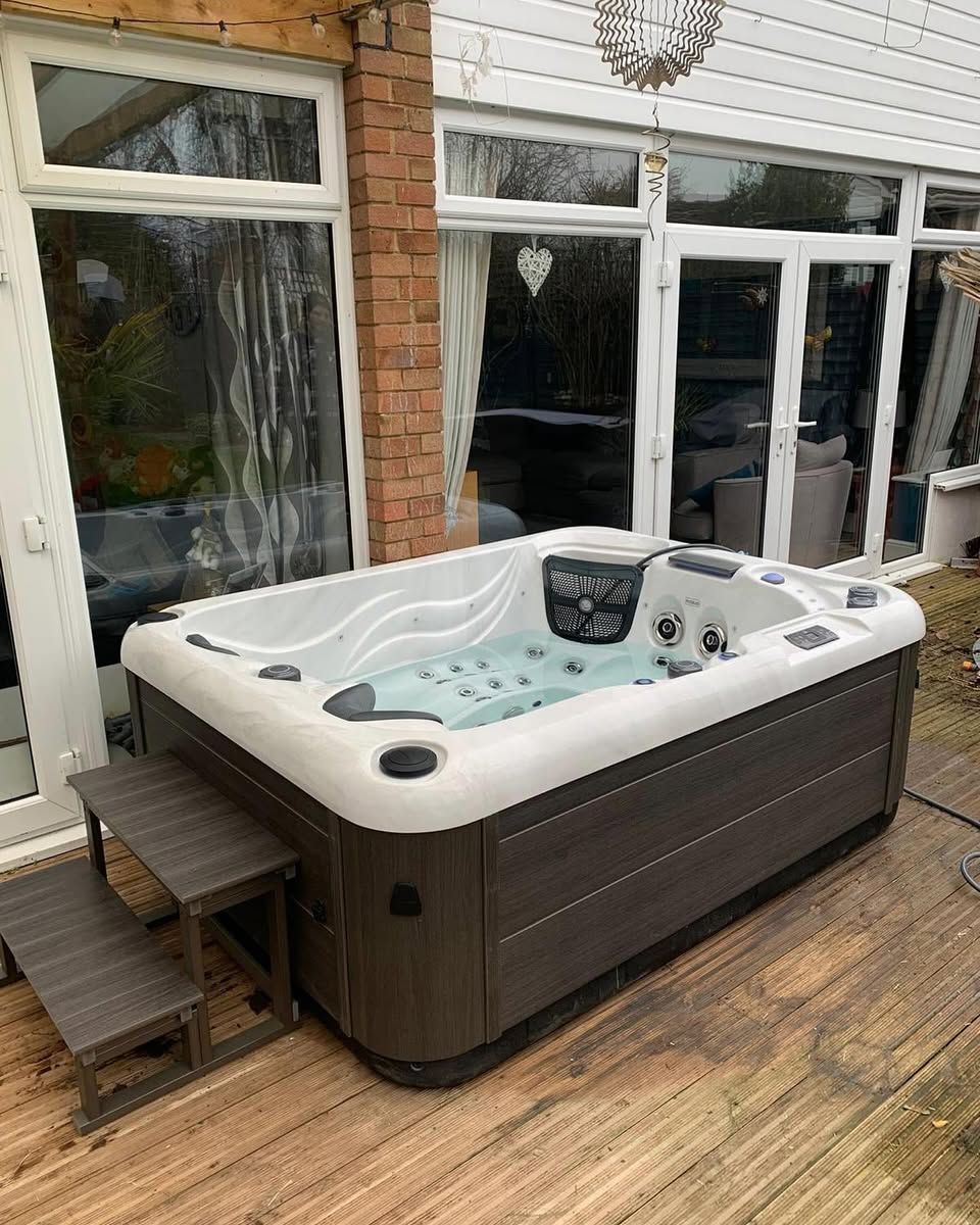 Hot tub on wooden deck, next to a glass-walled structure, with steps leading to it.