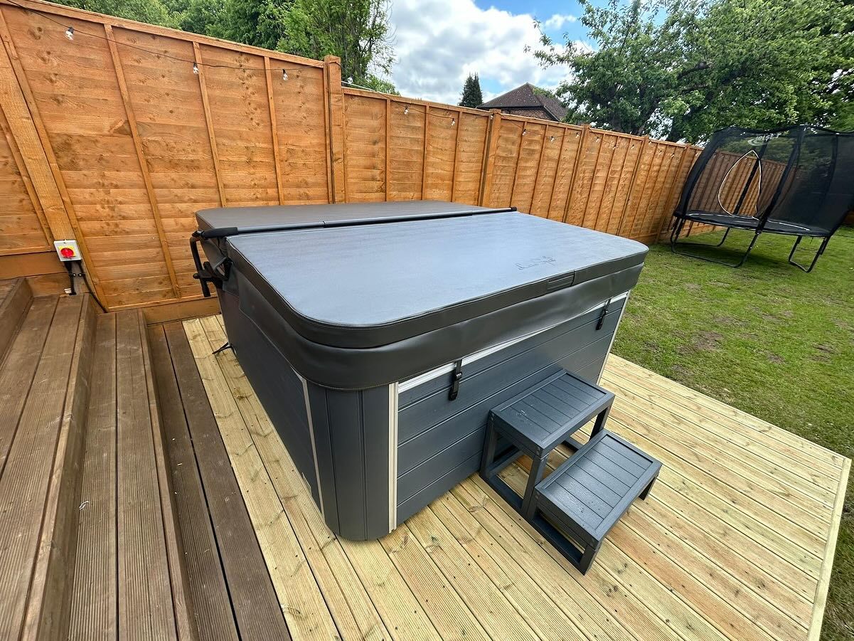 A hot tub with closed cover on a wooden deck, near a wooden fence and trampoline.