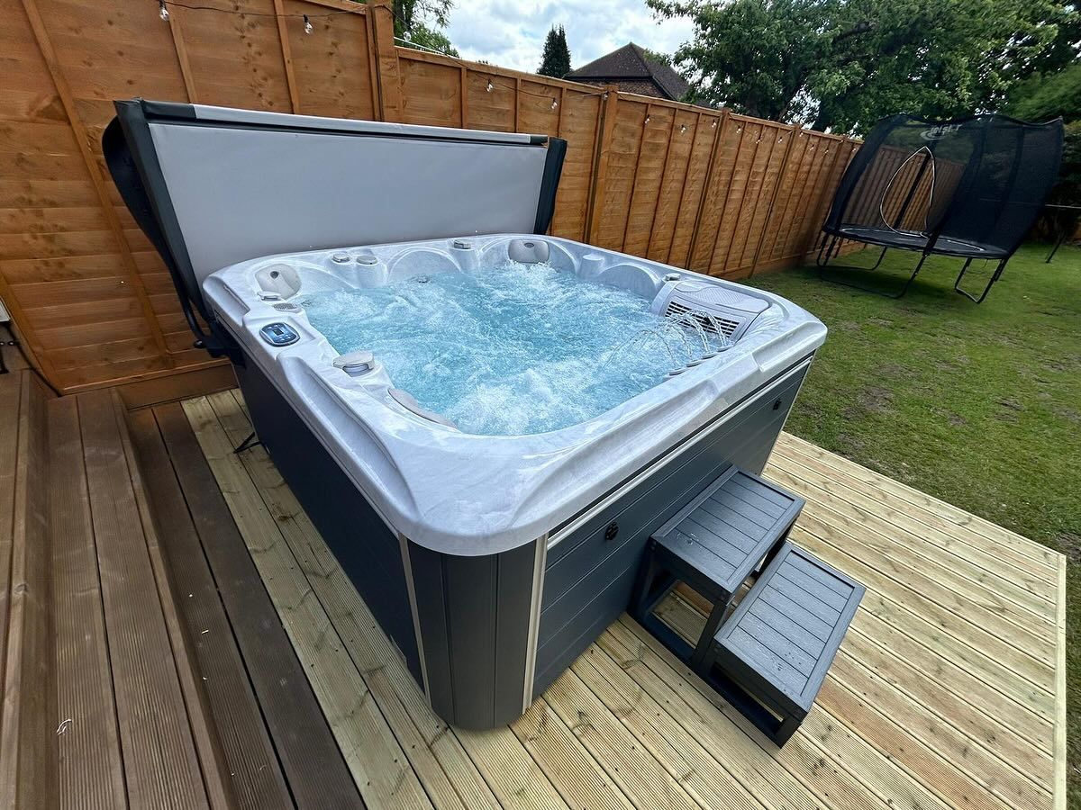 Hot tub on wooden deck, cover open, bubbles rising, steps to enter, fenced yard, cloudy sky.