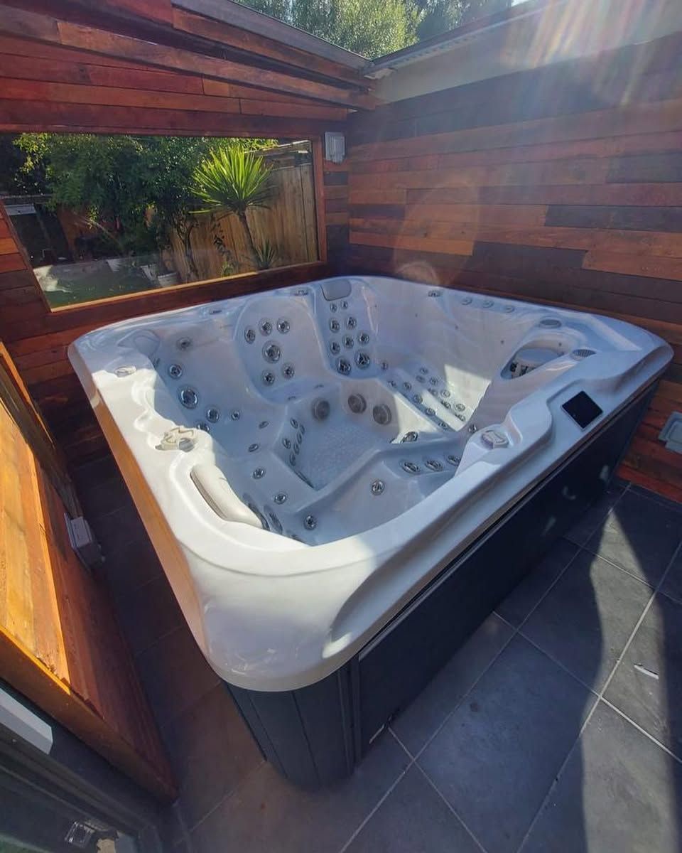 Hot tub surrounded by wooden structure, set on tiled patio with plants visible in background.