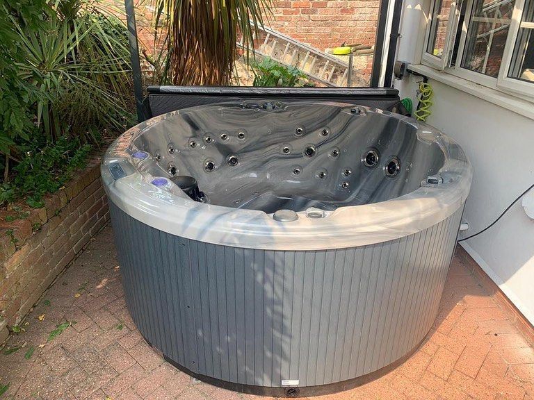 Round grey hot tub on a brick patio, in front of a white building with a window.