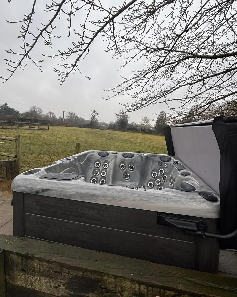 Hot tub with open cover, set on a wooden deck in front of a grassy field, under bare tree branches.
