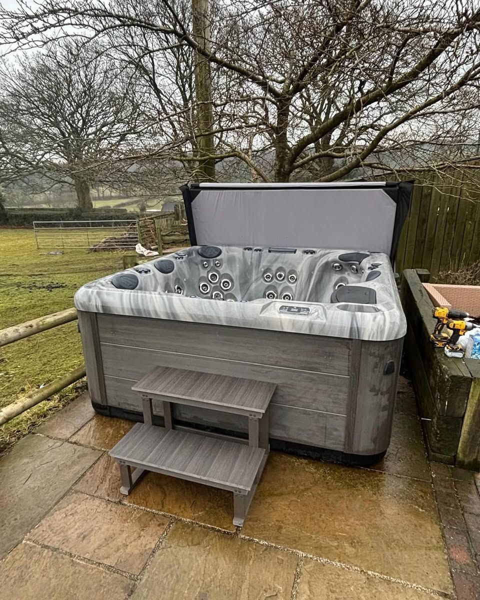 Hot tub with open gray cover, two-step stairs, and surrounding trees in an outdoor setting.