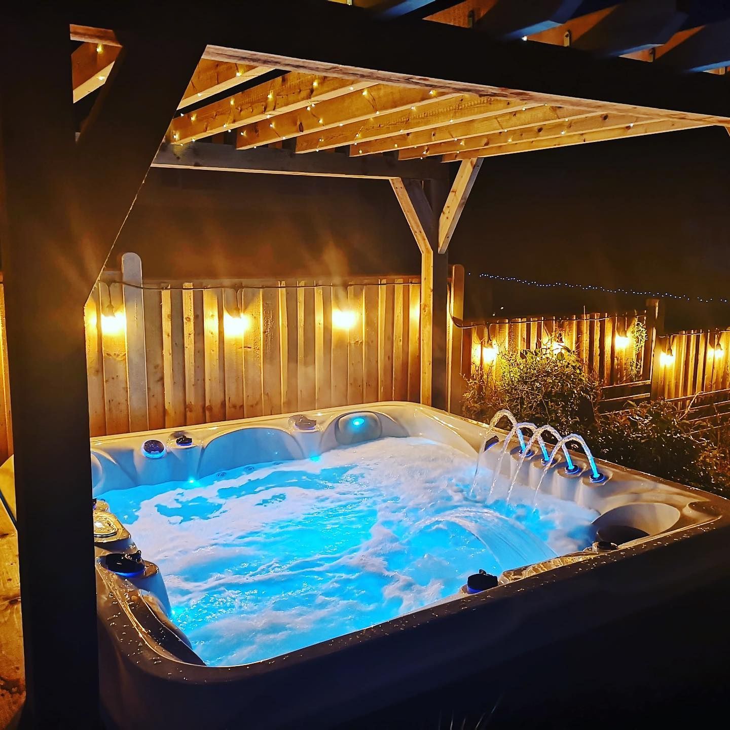 Lit hot tub under a wooden pergola at night, with illuminated jets, surrounded by string lights.