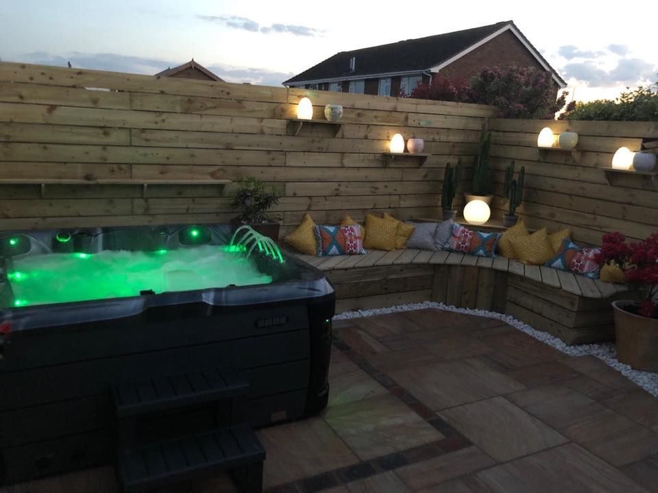 Hot tub on a patio with built-in seating, wooden fence with lights. Green water, pillows, evening setting.