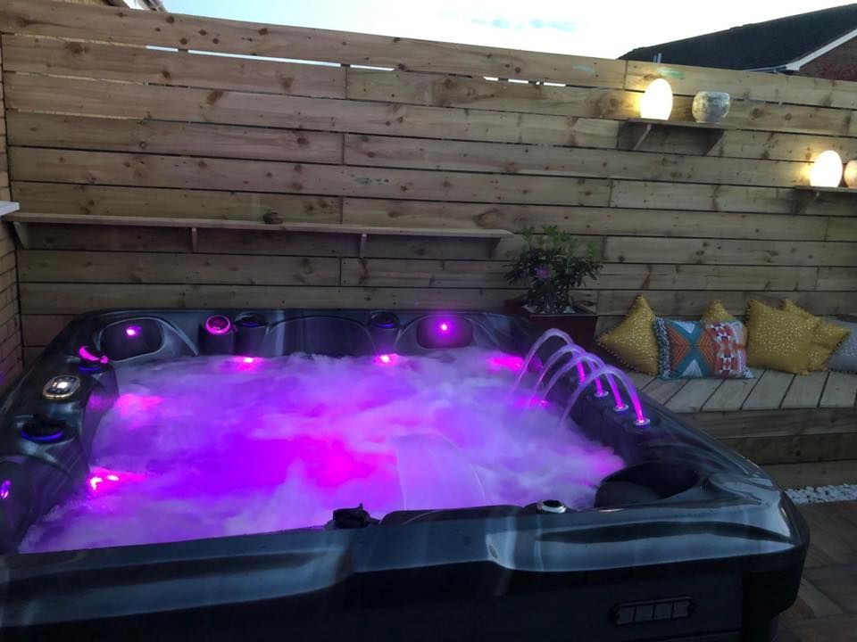 Lit-up hot tub with purple lights and bubbles, in a wooden-fenced outdoor setting. Cushions on a bench nearby.