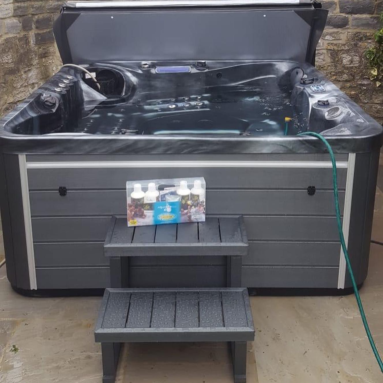 Hot tub with open cover, steps, chemicals, and hose in an outdoor setting. Gray and black.