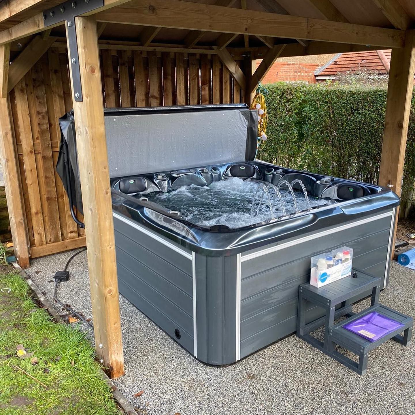 Hot tub under wooden gazebo, bubbling with open cover; steps to access, on gravel.