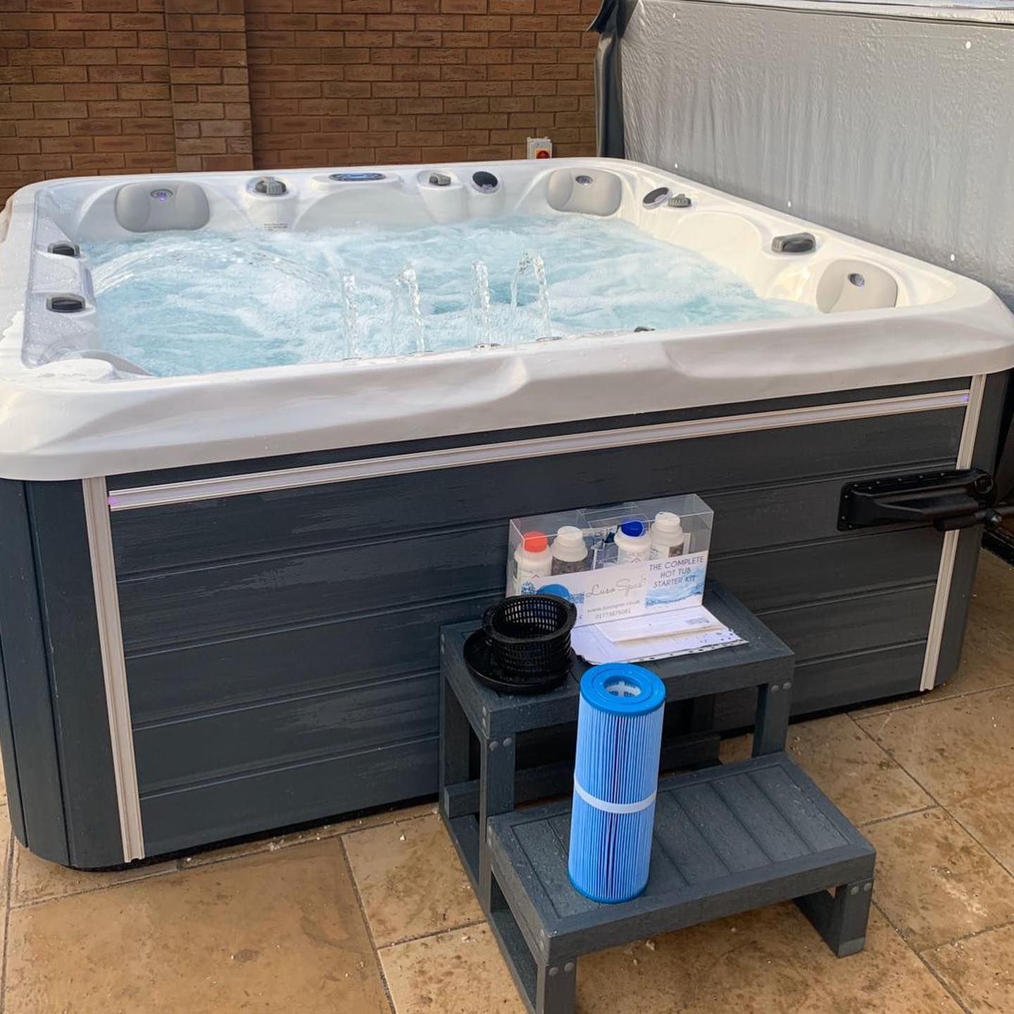 A filled hot tub with jets on, steps and accessories beside it on a patio.