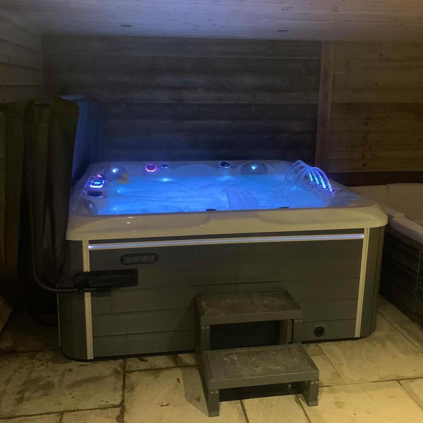 Hot tub with blue lights, steps in front. Grey and white tones, wooden background.