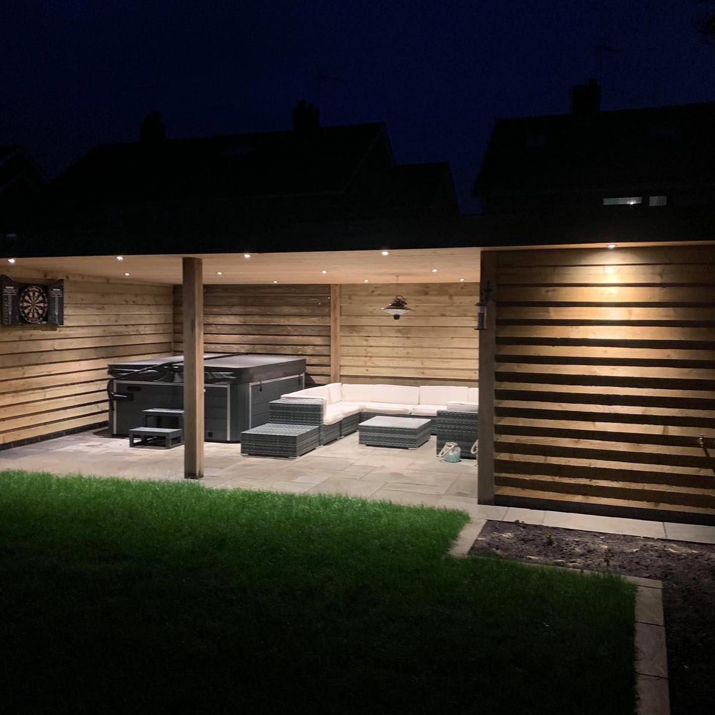 Outdoor wooden patio with a hot tub and seating, illuminated by lights at night.