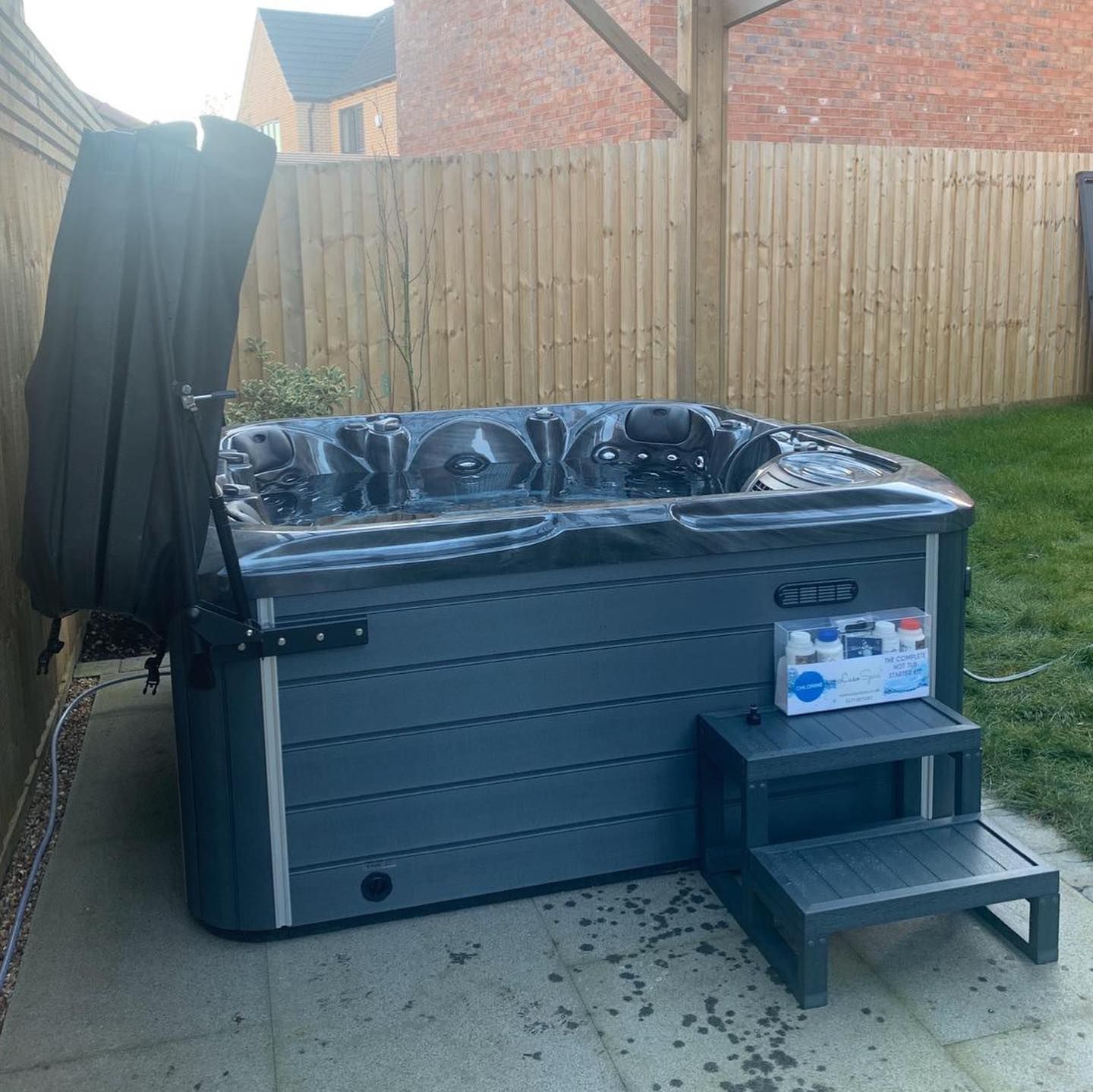 Hot tub with open cover, grey siding, black steps, in a backyard.