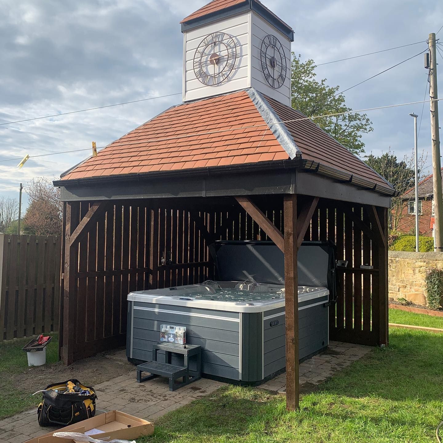Hot tub in gray gazebo with clock tower on a brick pathway, surrounded by a wooden fence on a lawn.