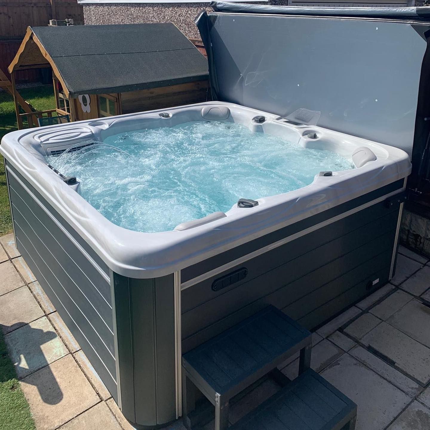 Bubbling hot tub with partially open gray cover, set on a patio, with a step.