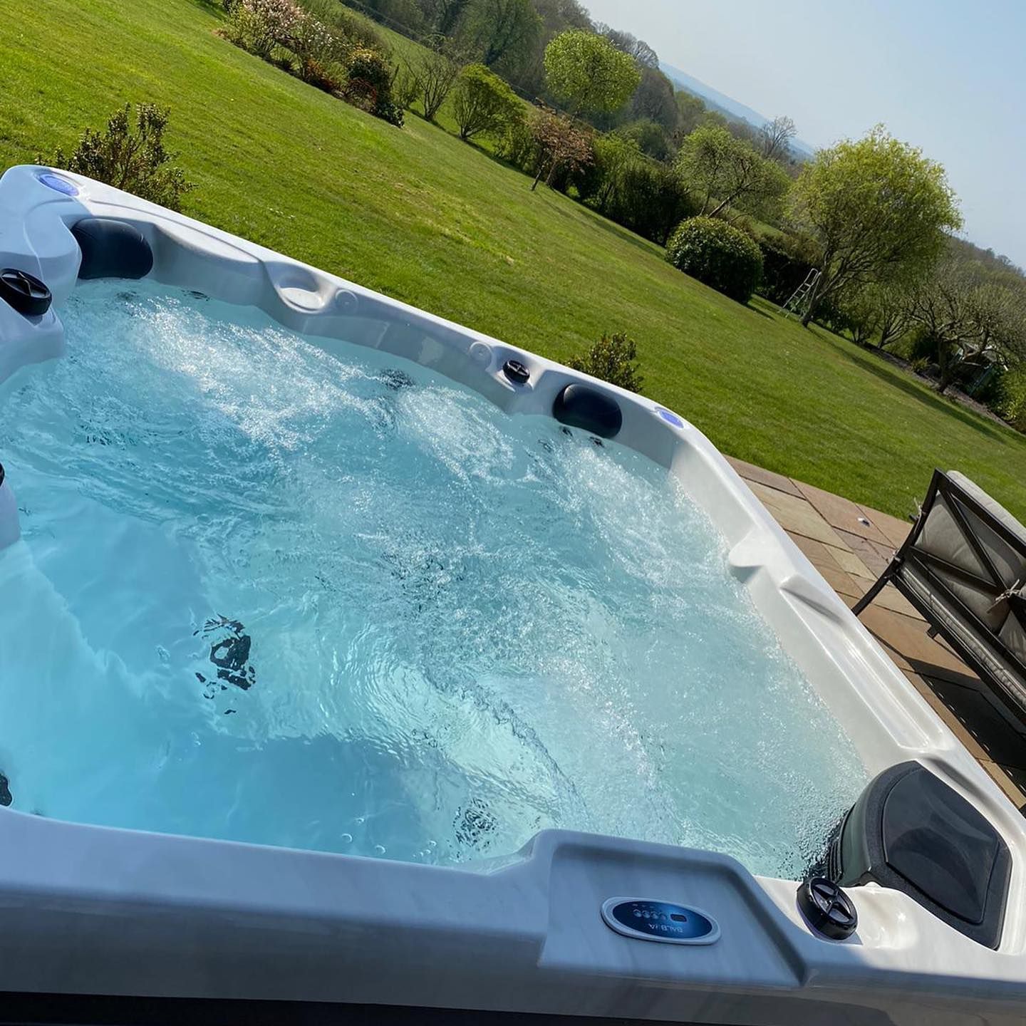 Hot tub with clear water, outdoors on a sunny day. Green hillside in background.