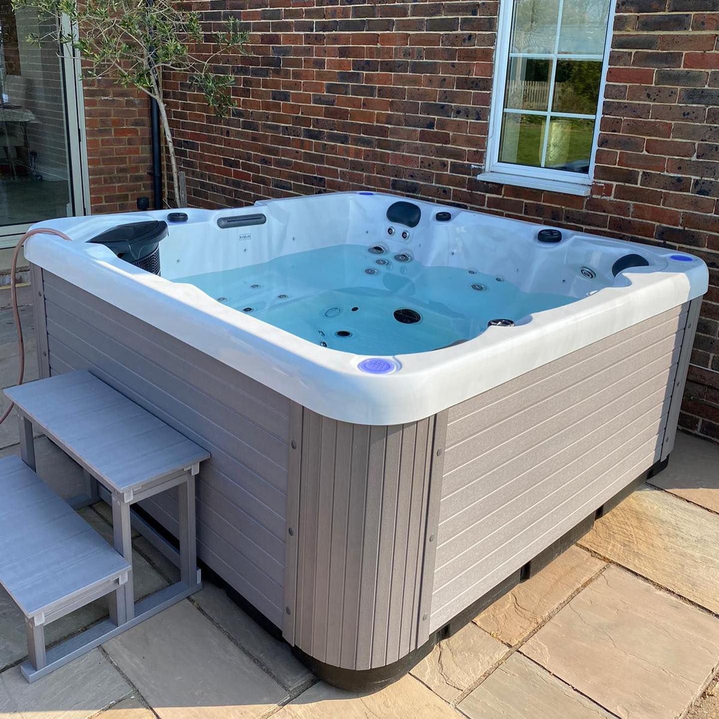 A square hot tub with water, surrounded by gray siding, and steps on a brick patio.