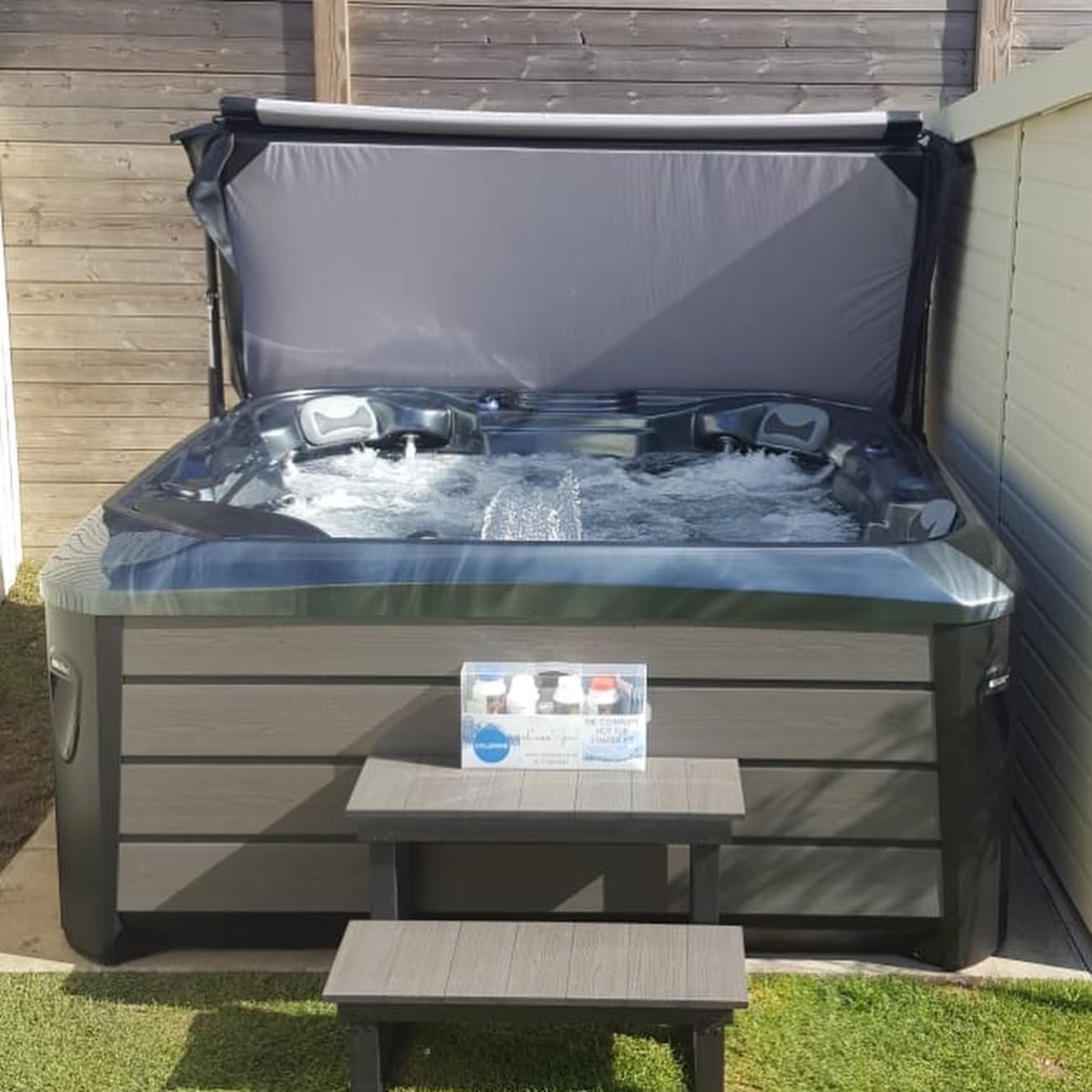 Hot tub with gray cover open, gray steps, box of supplies. Outdoors, daytime.