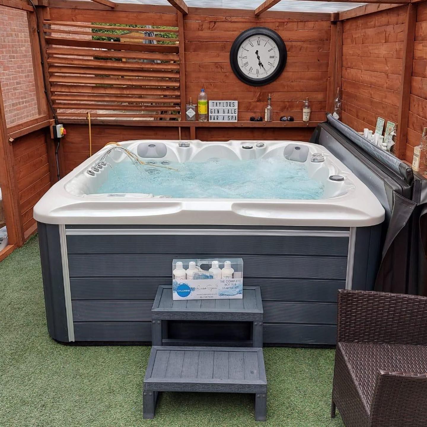 Hot tub in a wooden enclosure with a clock, bottles, and a step stool.