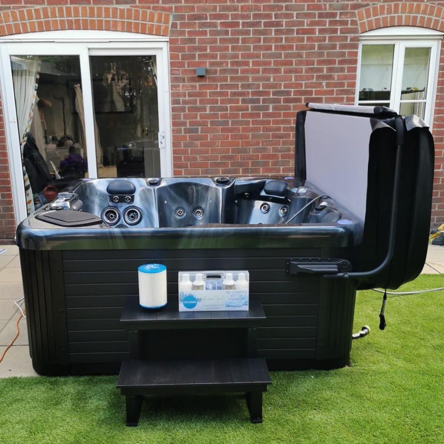 Black hot tub with open cover, next to steps, filter, and chemicals, on a lawn in front of a brick wall.