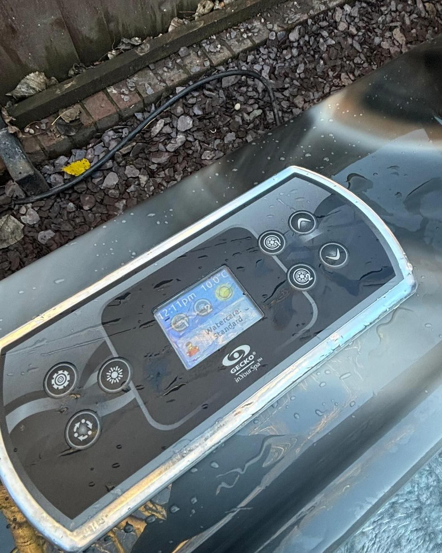 Close-up of a black and gray hot tub control panel with buttons and a small blue display outdoors.