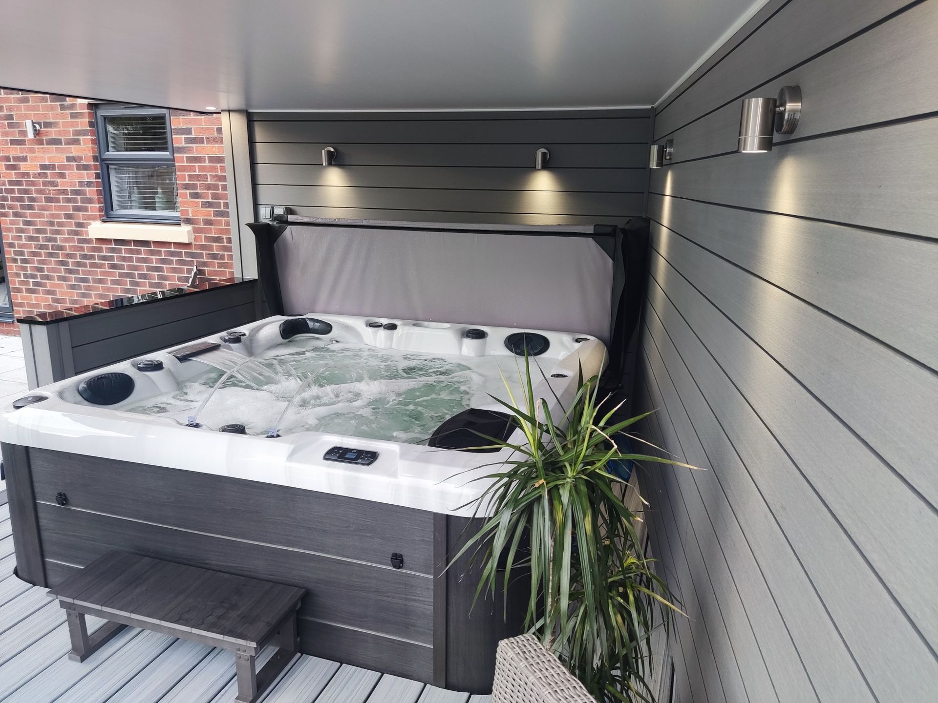 Hot tub in an enclosed deck area, gray siding and lighting. Plant next to tub, brick building in background.