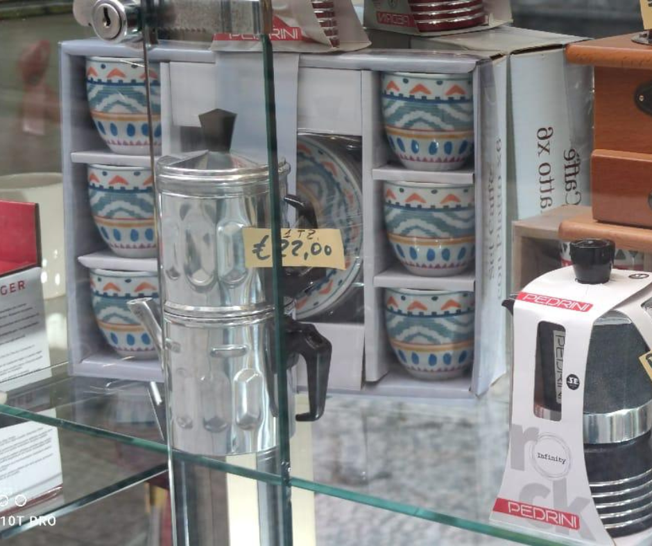 Uno scaffale di vetro con tazze decorate impilate e una piccola tazza di metallo con l'etichetta €2,20 in una vetrina di un negozio.