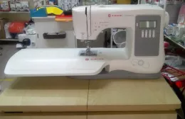 Una macchina da cucire bianca su un tavolo di legno in una stanza disordinata, con tessuti e accessori sullo sfondo.
