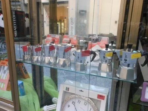 In una vetrina, macchine da caffè argentate e articoli con confezioni rosse sono esposti dietro una teca di vetro.