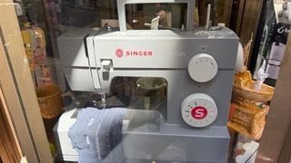 Una macchina da cucire Singer grigia esposta in una vetrina, con tessuti e cestini intorno.