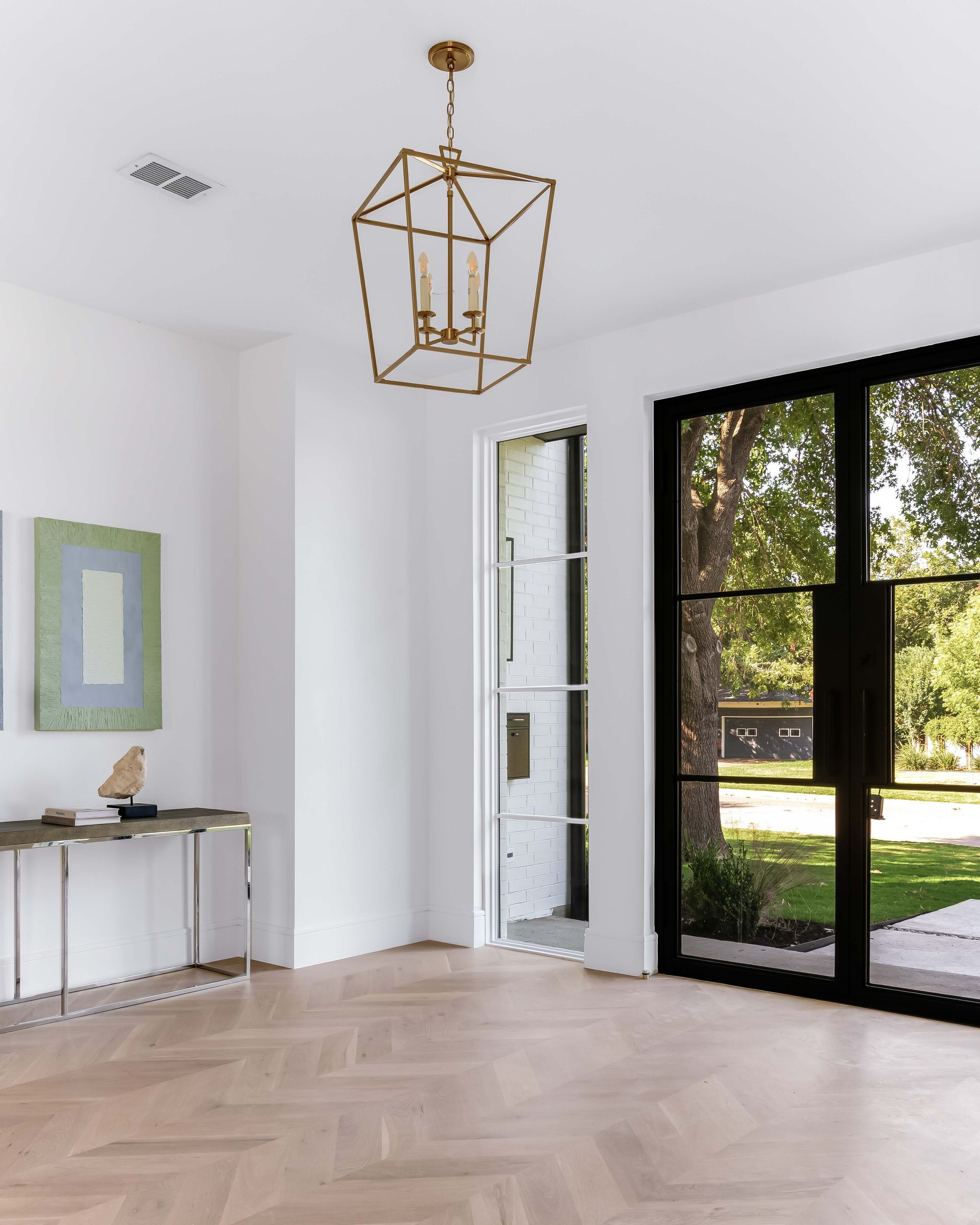 Gold geometric chandelier hangs in a bright room with light wood floors and black-framed glass doors.