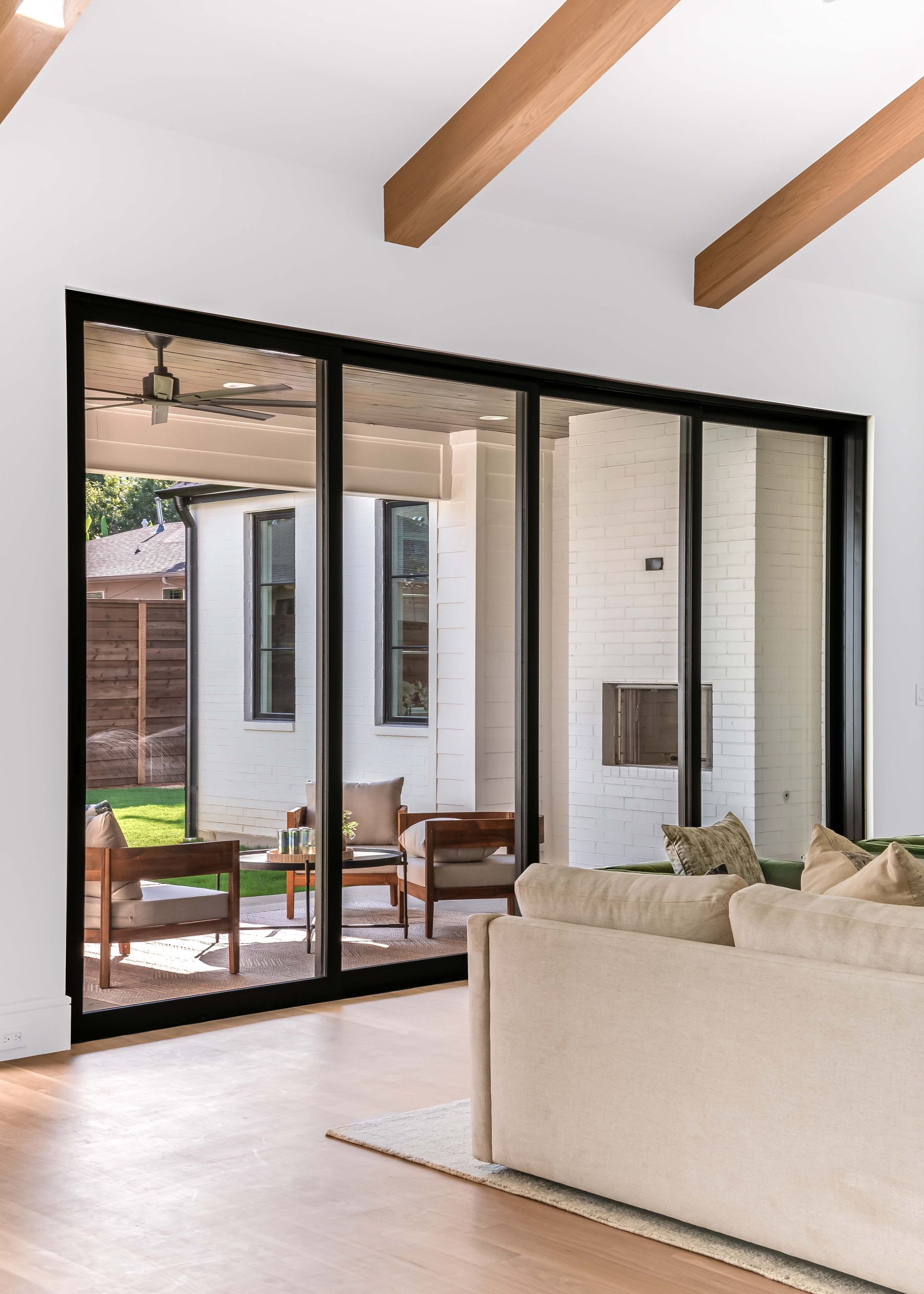 Living room with large black-framed sliding glass doors, leading to a patio with seating. Beige sofa and wooden beams.