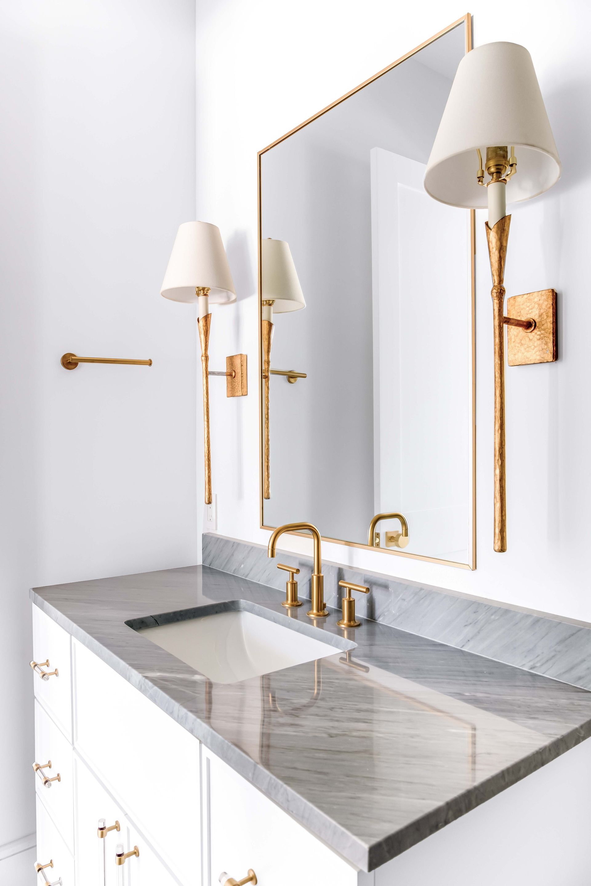 White bathroom with a gray countertop, gold fixtures, mirror, and sconces.