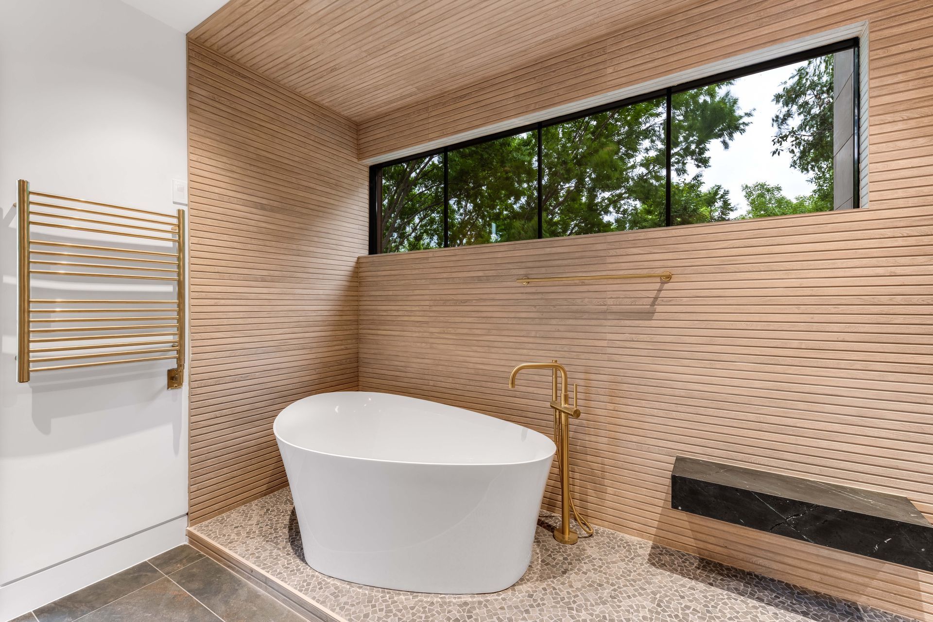 Modern bathroom with a white tub, gold fixtures, and wood-paneled walls and ceiling, with a view of green trees outside a window.