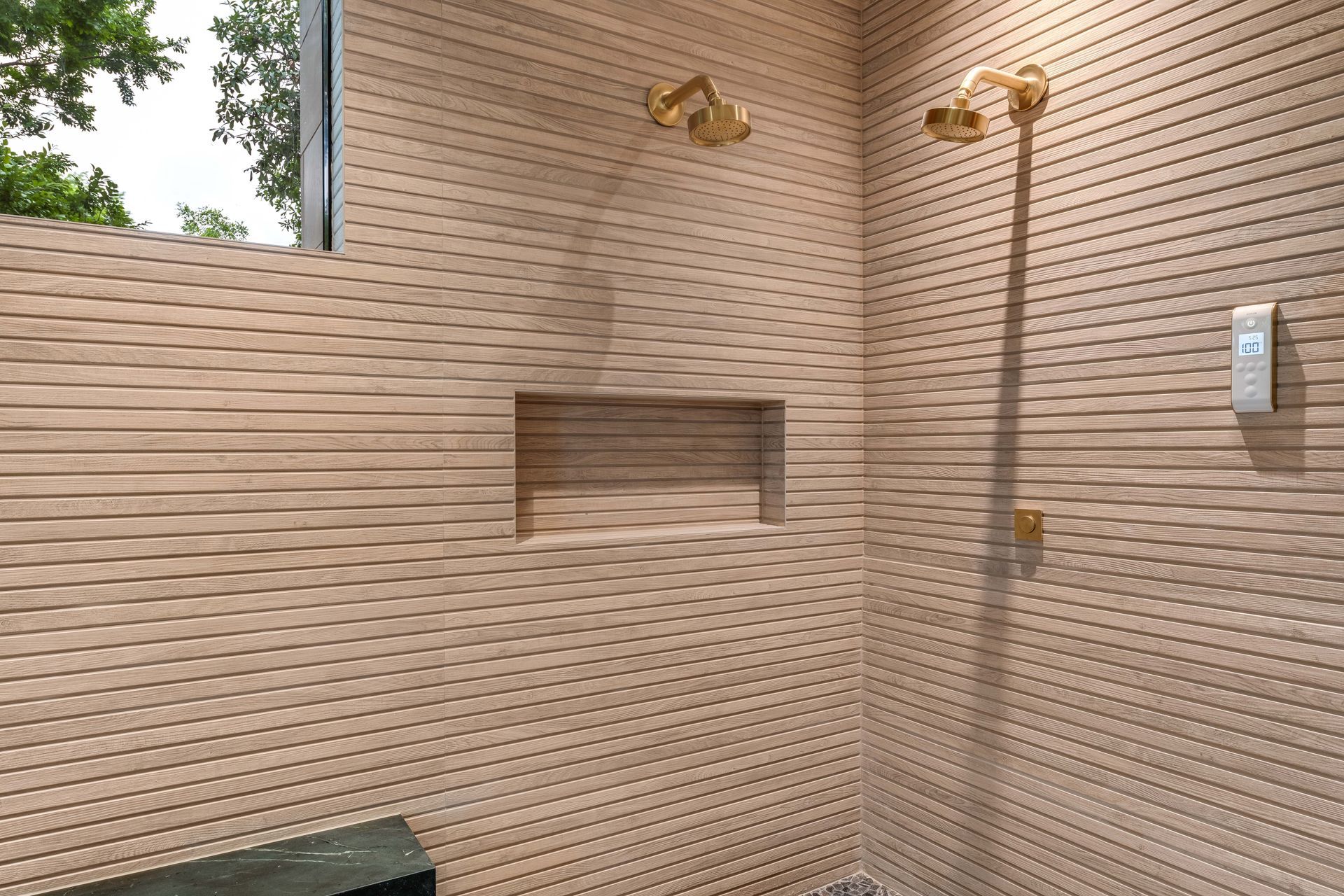 Shower with textured tan tiled walls, gold shower heads, and a recessed shelf.