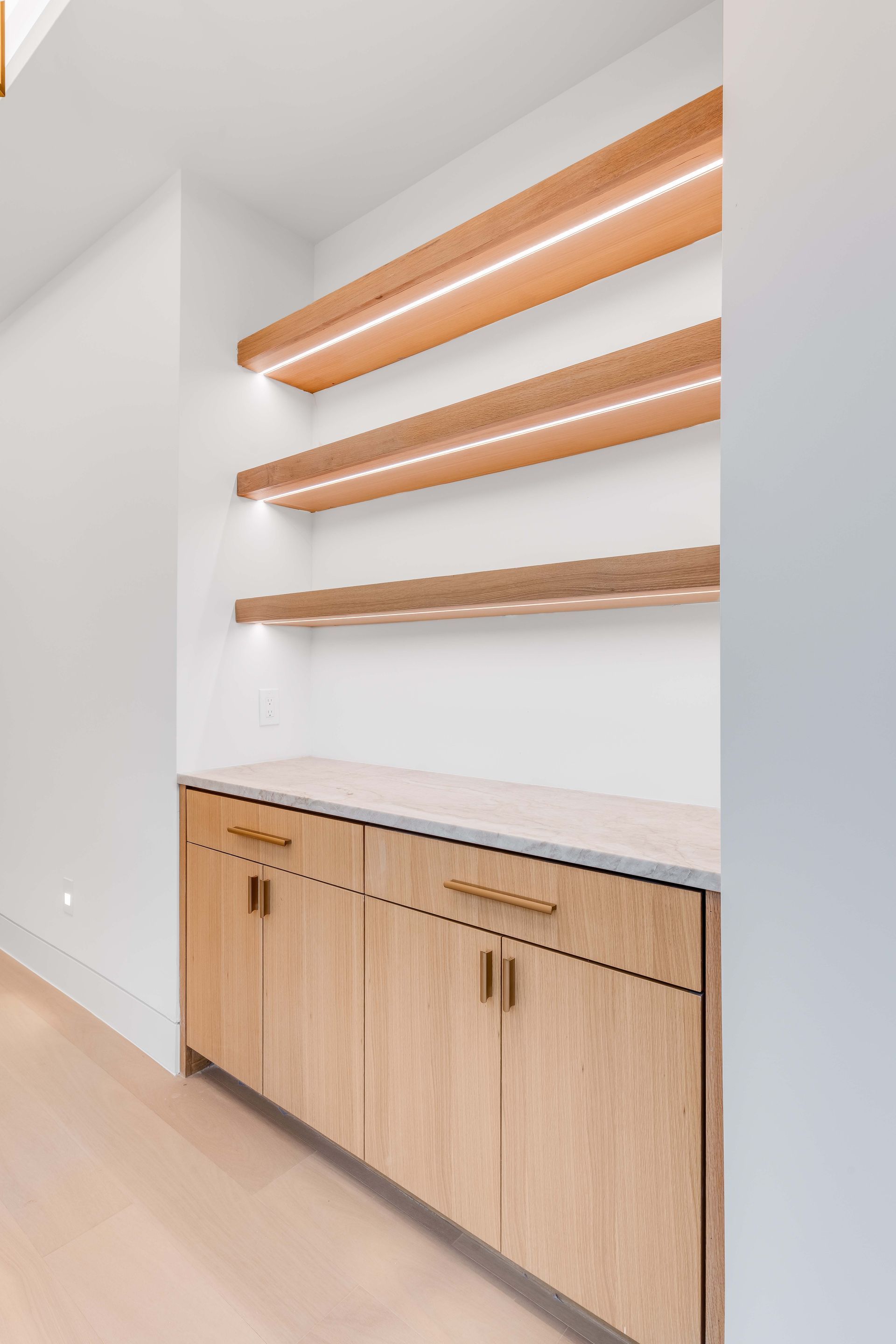 Built-in wooden shelving with warm lighting above lower cabinets; light wood and marble countertop.