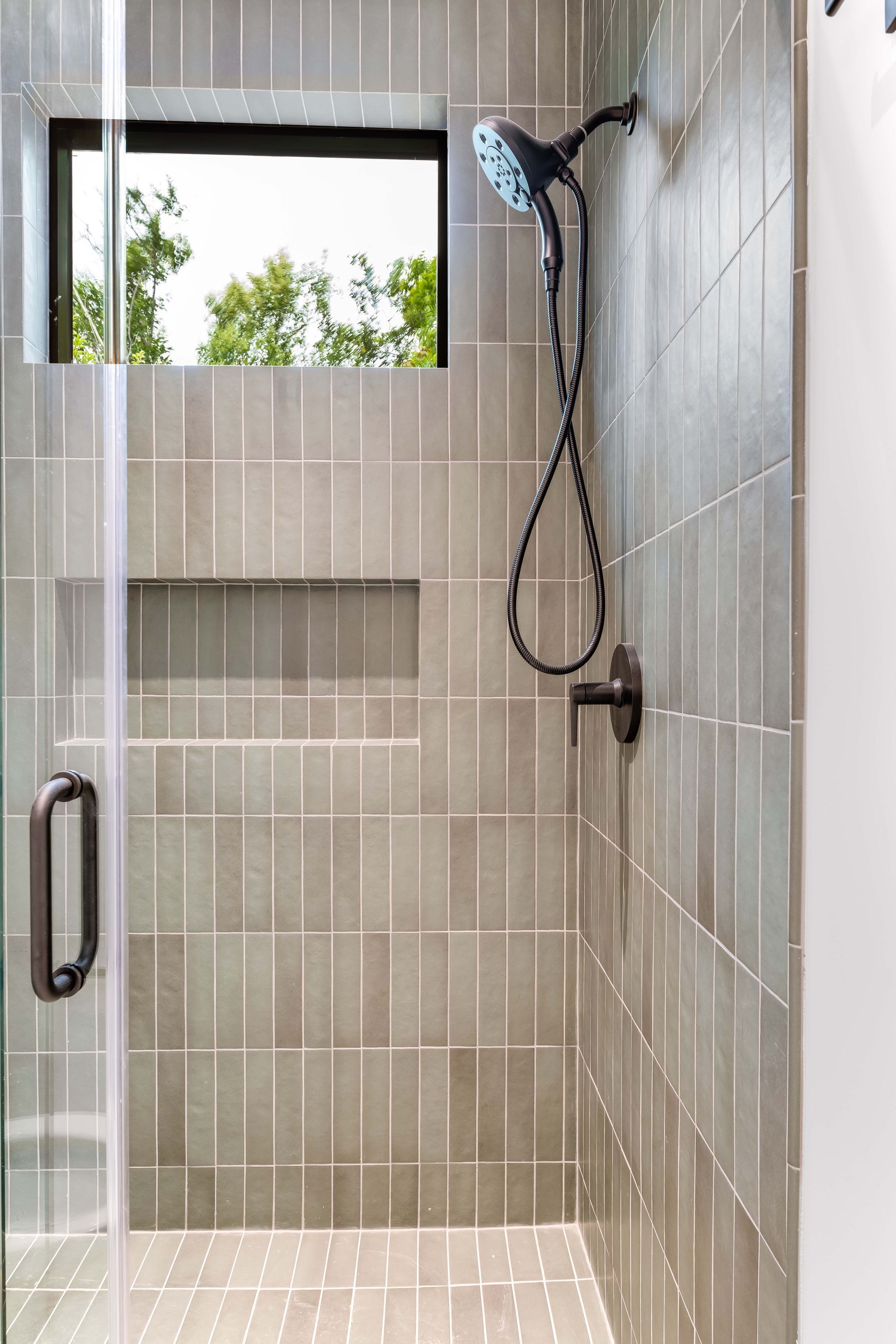 Shower with gray tiled walls, black shower fixtures, a window, and a glass door.