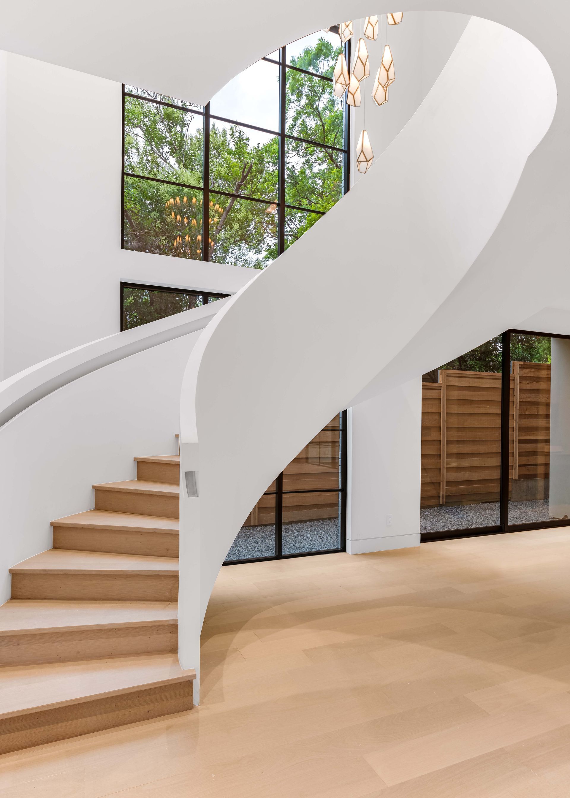 Curving white staircase with wooden steps, leading to a large window with a view of trees.