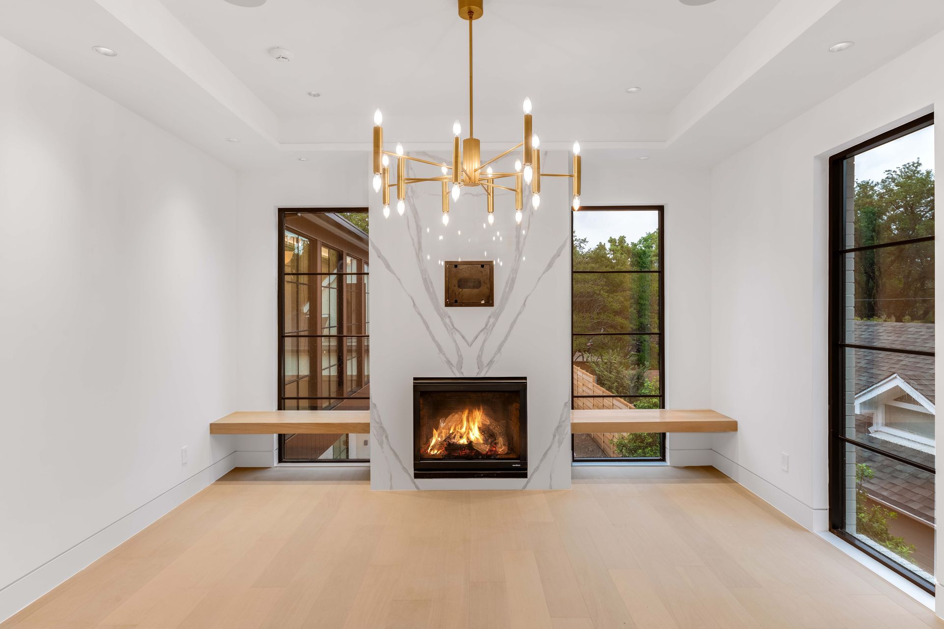 Modern living room with fireplace, wood floors, large windows, gold chandelier, and marble accent wall.