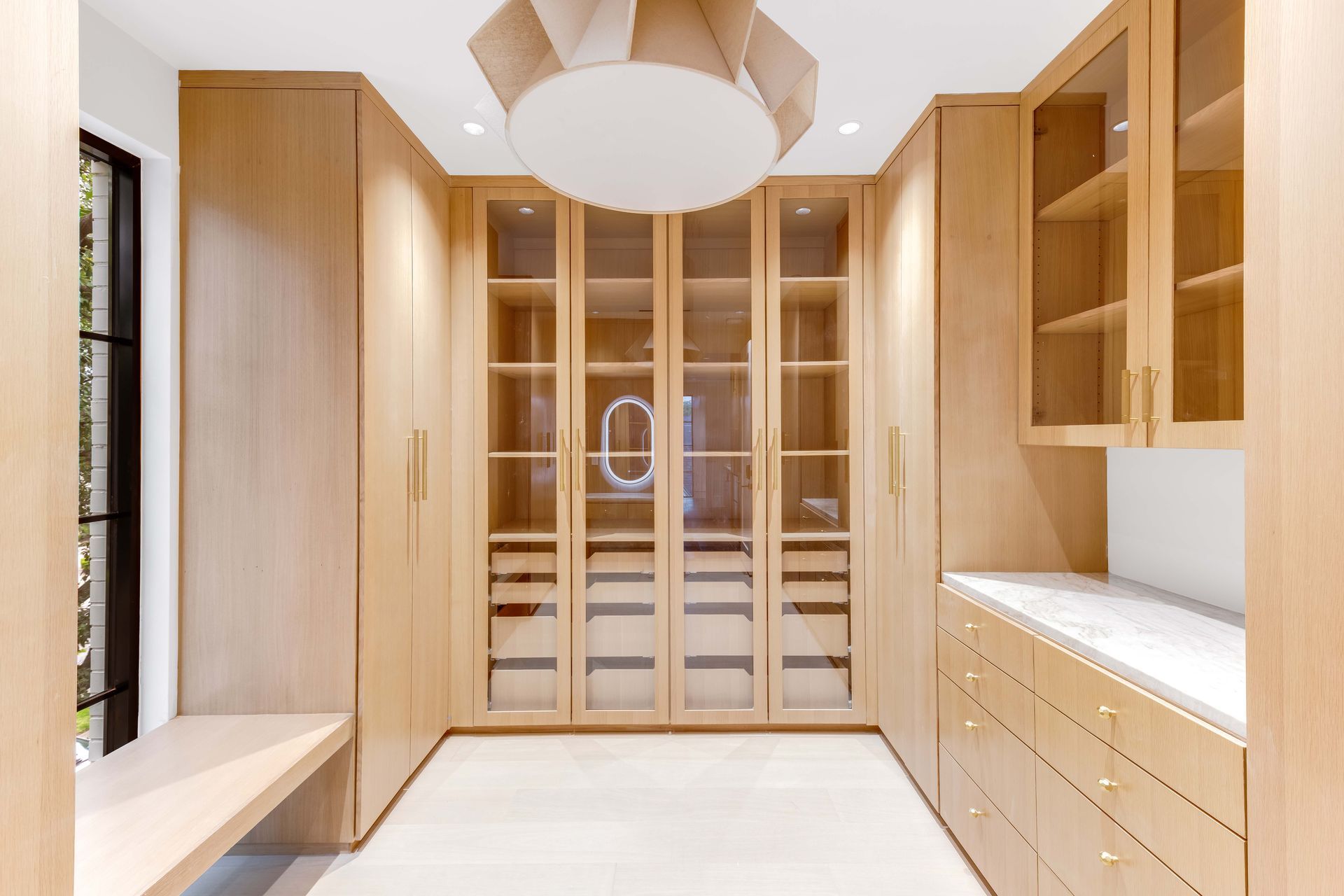Light-wood walk-in closet with glass-front cabinets, drawers, and a vanity area.