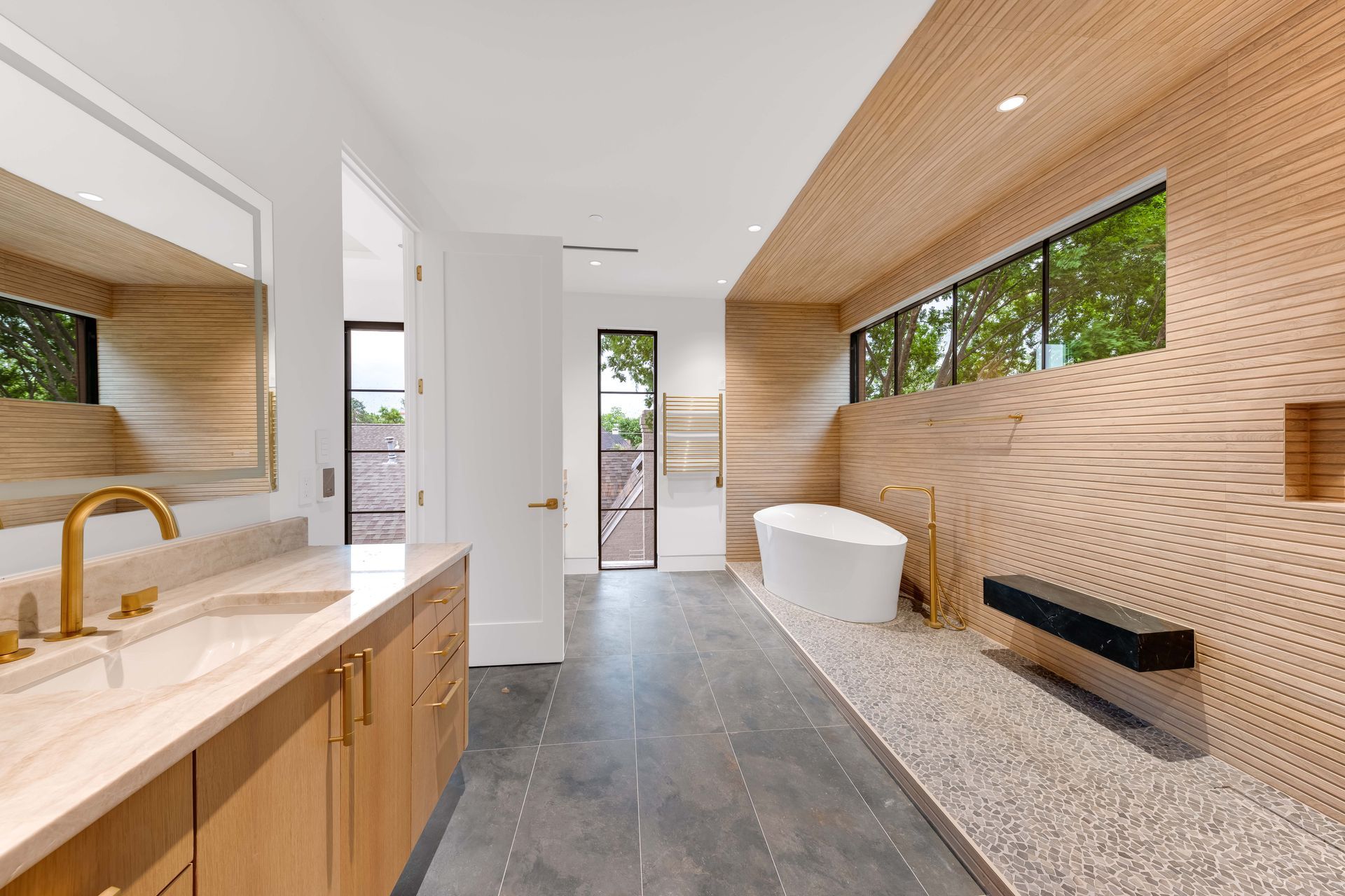 Modern bathroom with wood accents, stone flooring, soaking tub, and vanity with gold fixtures.