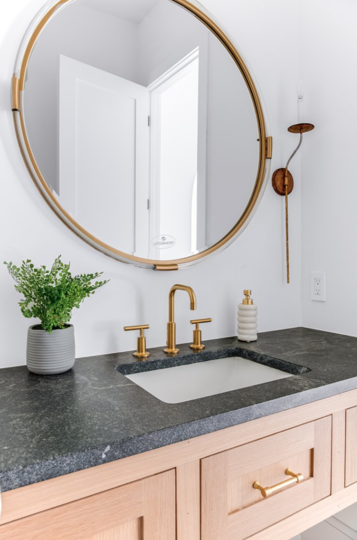 Bathroom with gold accents: round mirror, faucet, sconce; black countertop, light wood cabinets, potted plant.