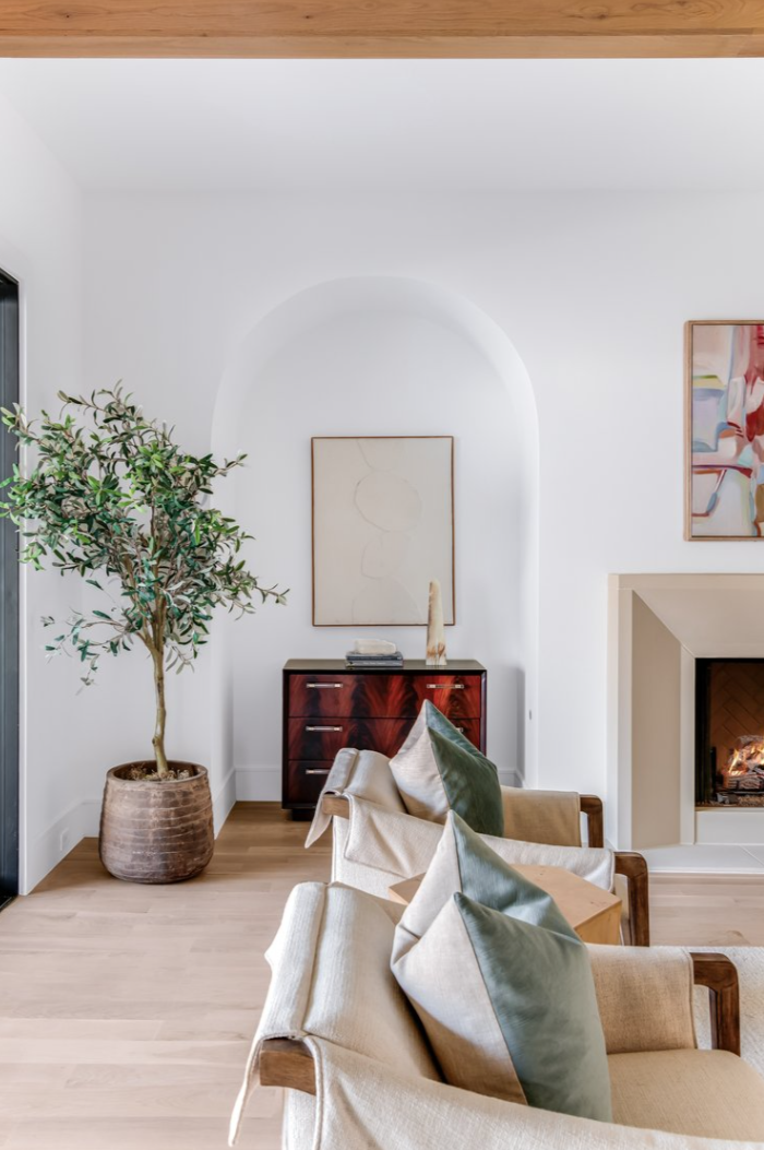 Living room: white walls, arched niche with art, olive tree, cream seating, fireplace.