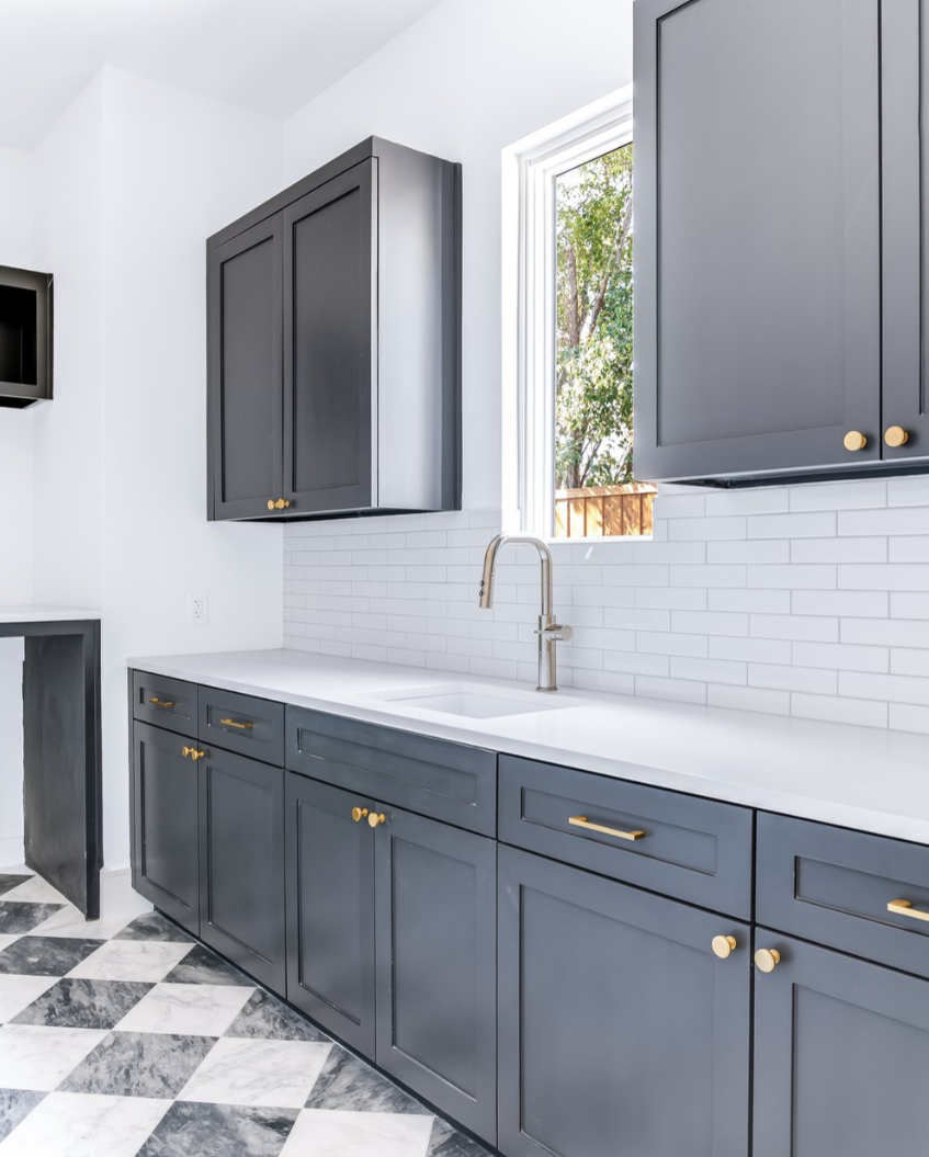 Dark gray cabinets with gold hardware in a kitchen with white countertop, subway tile backsplash, and checkered floor.