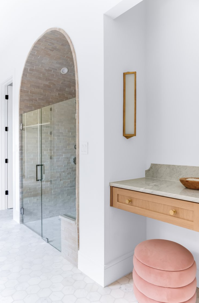 Bathroom with vanity and archway to shower; blush stool, gold accents, white walls, wood tones.